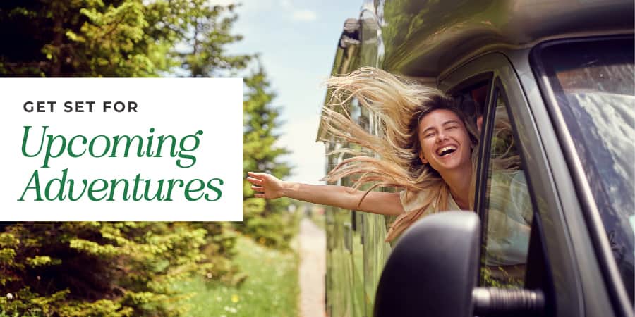 A woman half-way out the passenger side window of an RV enjoying the outdoor breeze. At the left, the words: get set for upcoming adventures.