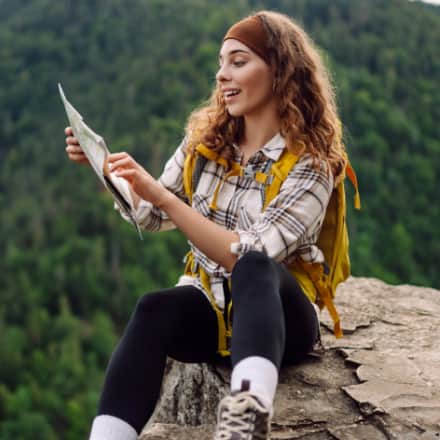 A smiling female hiker sitting on a rock looking at a map with mountains in the background.