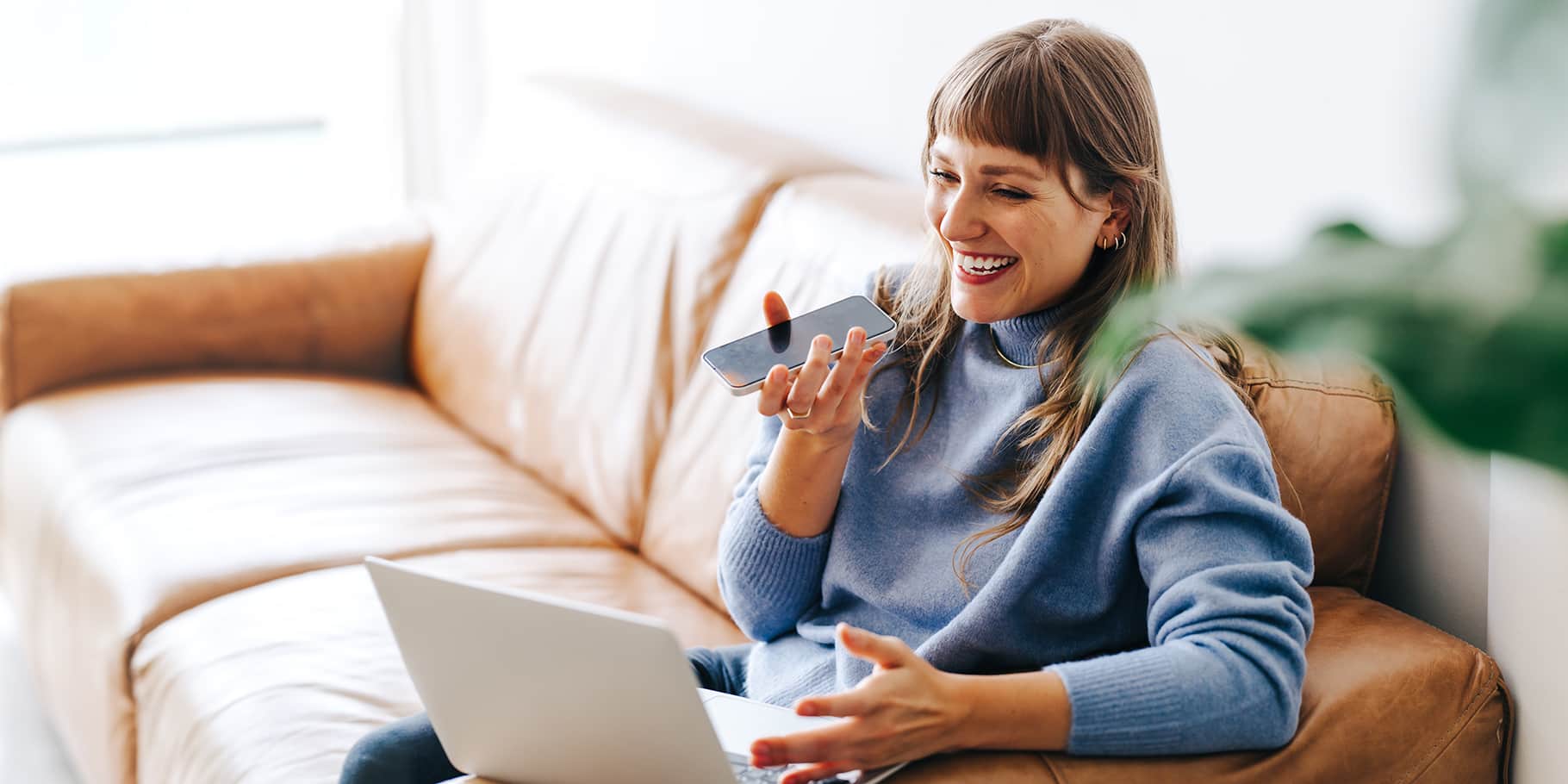 A woman sitting on a couch speaking using a mobile phone in speaker mode.