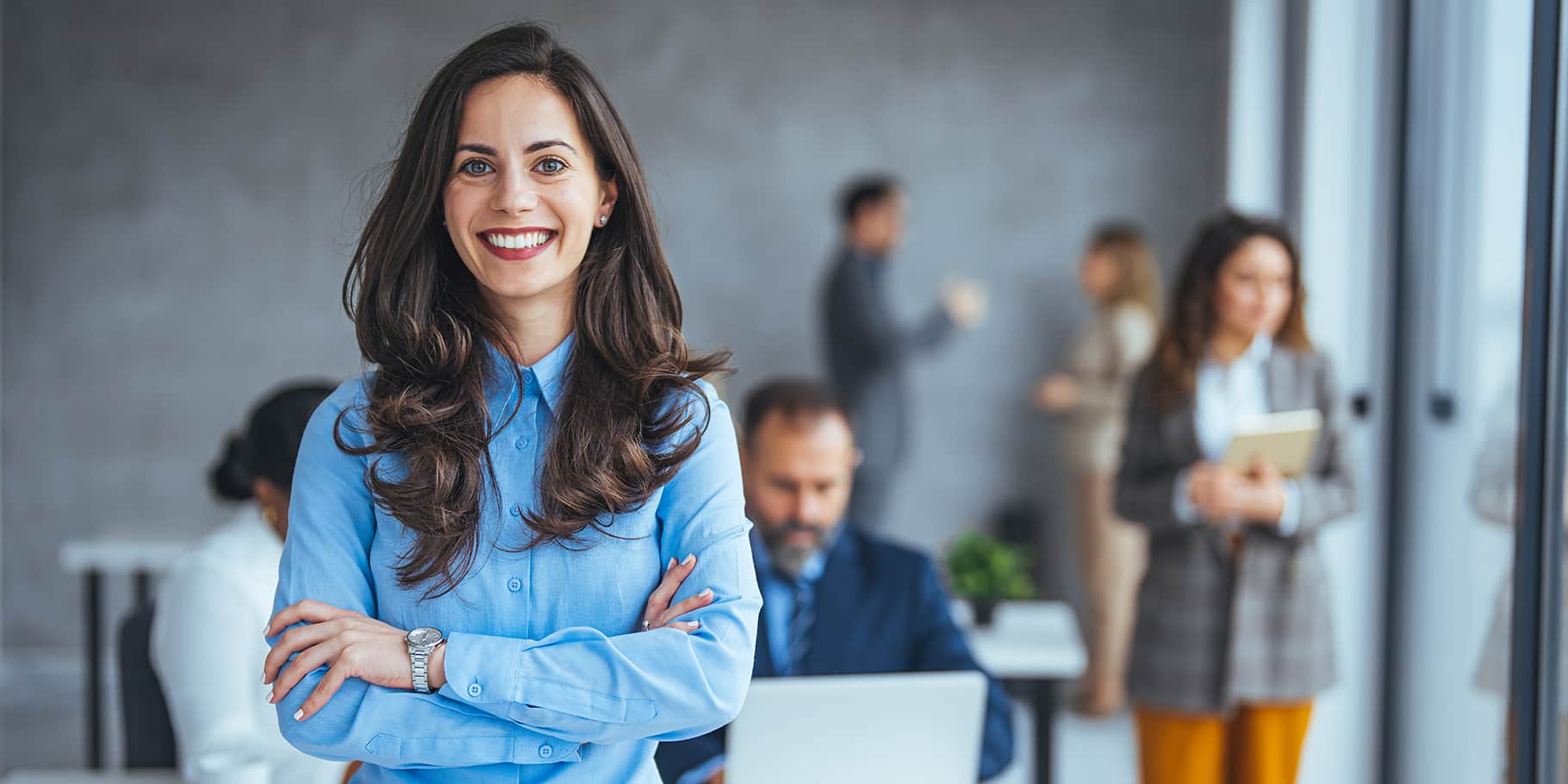 A smiling woman in an office setting with employees in the background.