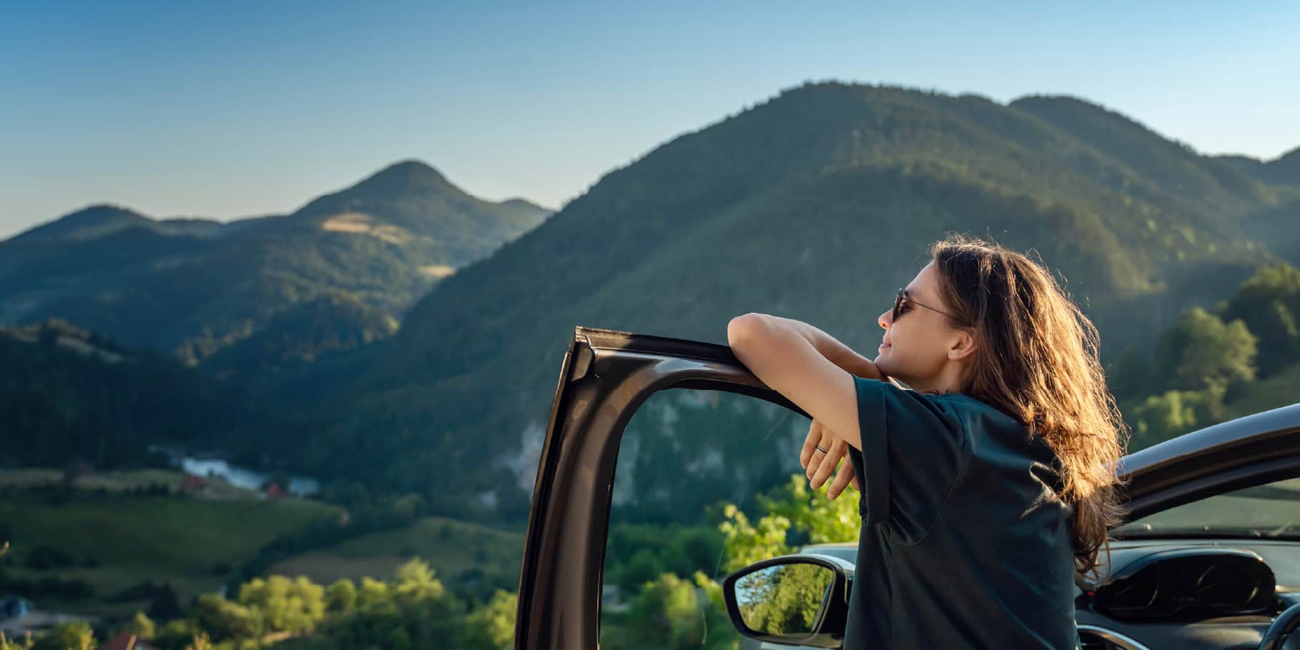 A woman leaning against her car window looking out into a beautiful mountain view.